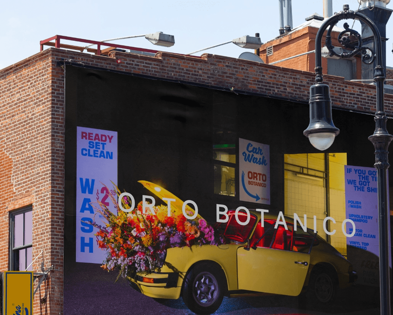 Giant billboard in Brooklyn on a brick wall for Orto Botanico, showing a 1970s carwash scene with a yellow Porsche opened and filled with colorful flowers; branding by Studio La Folie.